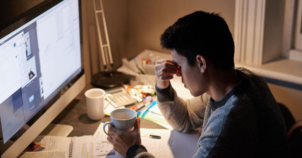 A stressed young man holding a coffee mug and looking at the desktop monitor.