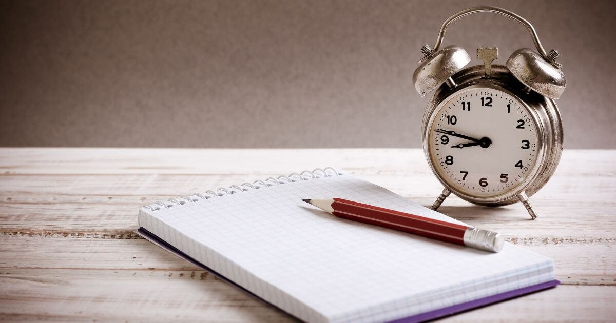 A traditional alarm clock, a pencil, and a notebook on a desk.