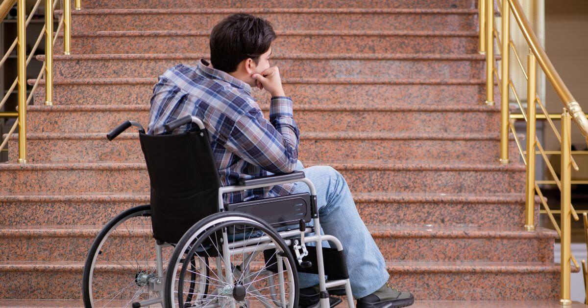 A young man sitting in a wheelchair, in front of an outdoor staircase. The knuckles of his hand rest on his cheek.
