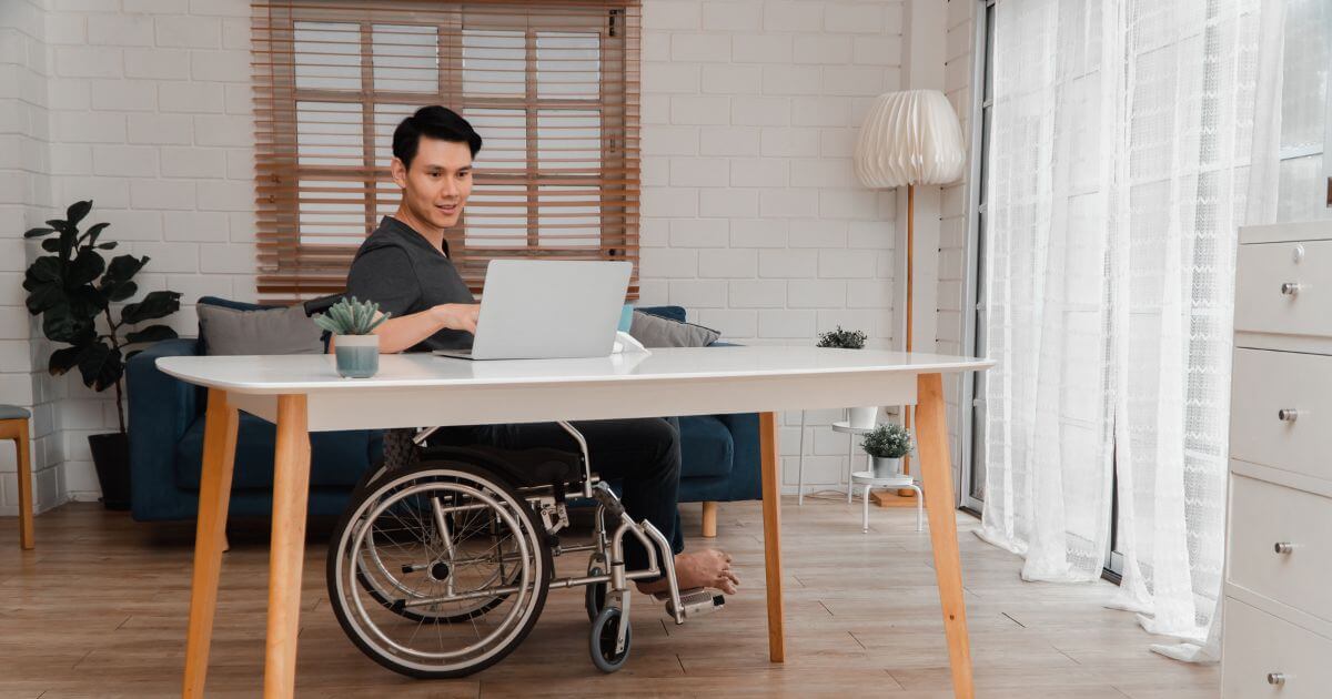 A smiling young man in wheelchair working on a laptop at his dining table.